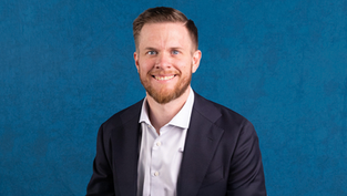 Smiling man in a suit and white shirt against a textured blue background. He has short hair and a beard. Mood is professional and friendly.
