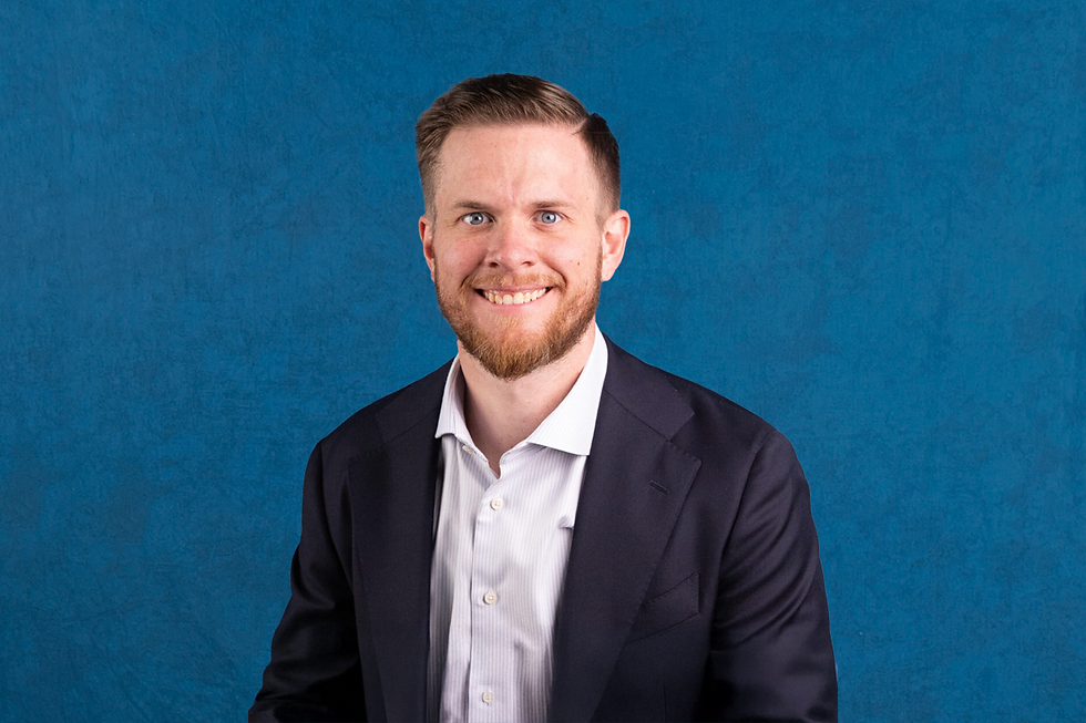 Smiling man in a suit and white shirt against a textured blue background. He has short hair and a beard. Mood is professional and friendly.
