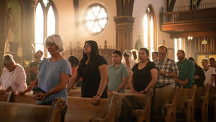 People standing and praying in a sunlit church with stained-glass windows. The mood is serene, with soft light highlighting wooden pews.