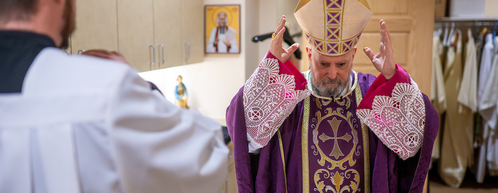 A bishop in ornate purple vestments and mitre gestures solemnly in a room, facing a priest in white. Religious decor in the background.