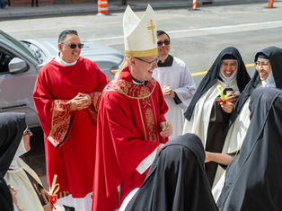 Bishop in red vestments and mitre smiles with nuns and clergy on a sunny street, exchanging palm crosses.