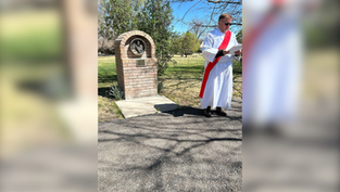 A person in a white robe with a red sash reads a document near a brick monument outdoors on a sunny day, with trees and grass in the background.