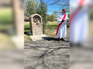 A person in a white robe with a red sash reads a document near a brick monument outdoors on a sunny day, with trees and grass in the background.