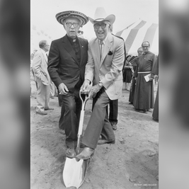 Dos hombres con sombrero posan sobre una pala en una ceremonia de inauguración, con carpas y clérigos detrás.