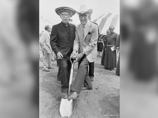 Dos hombres con sombrero posan sobre una pala en una ceremonia de inauguración, con carpas y clérigos detrás.