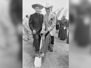 Dos hombres con sombrero posan sobre una pala en una ceremonia de inauguración, con carpas y clérigos detrás.
