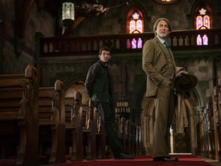 Two men stand in a dimly lit church with stained glass windows. One in a suit holds a hat, looking serious. Wooden pews line the aisle.