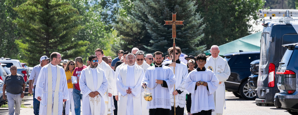Clergy in white robes lead an outdoor religious procession with a cross and incense, walking along a tree-lined street.