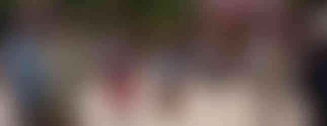 Runner in red shorts and a bull mascot approach the finish line at an outdoor event. Bystanders cheer under colorful flags.