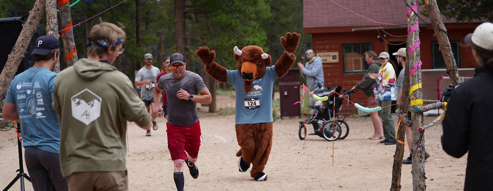 Runner in red shorts and a bull mascot approach the finish line at an outdoor event. Bystanders cheer under colorful flags.