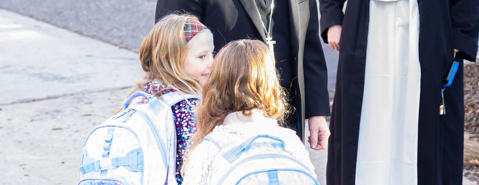 Two schoolgirls with backpacks labeled Caroline and Eleanor stand with a bishop and nun. Kids in the background. Bright, sunny day.