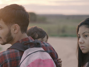 A man with a backpack carries a child, standing beside a woman. All wear plaid shirts in a serene rural landscape at sunset, looking thoughtful.