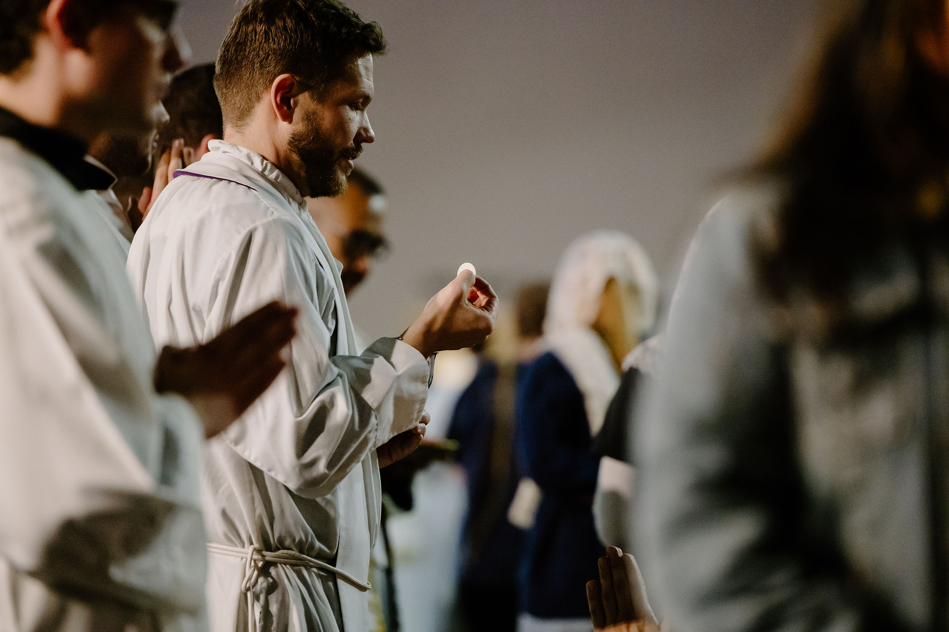 Priest in white robe holding the Eucharist in church setting, standing among other people, with a solemn atmosphere.