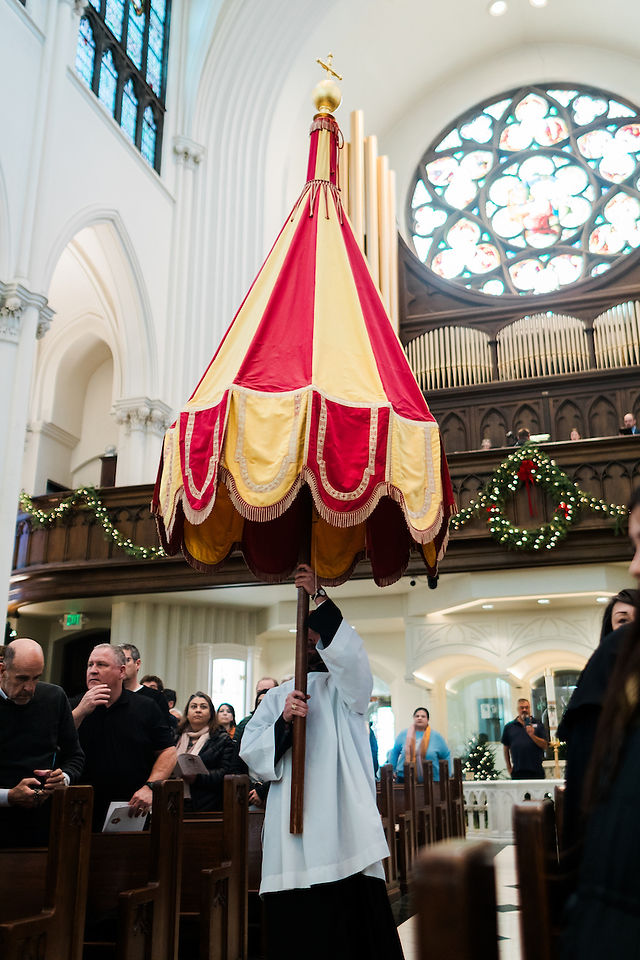 Person in a white robe holds a red and yellow canopy in a church with stained glass and greenery. Observers watch the procession.