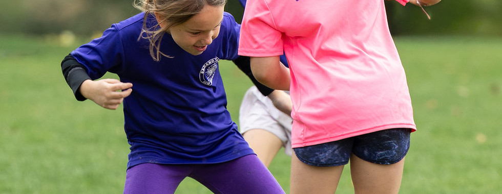 Children playing soccer on a grassy field, one in a pink VARSITY CATHOLIC shirt battling for the ball, trees blurred behind.