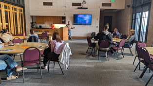 A speaker presents in a conference room with round tables. Attendees listen attentively. Screen displays "VIRTUS: Excellence Builds Trust."