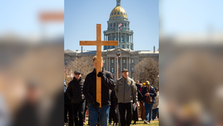 A man carries a large wooden cross in front of a crowd by a domed building. Clear sky and flags in the background. The mood is solemn.