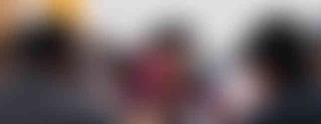 Man in glasses and maroon shirt gives thumbs-up at a table with others in a casual setting. Water bottle and flowers are on table.