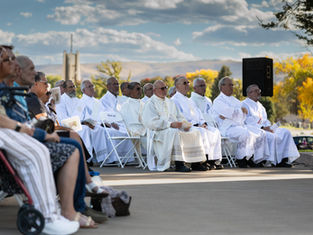 Clergy in white robes sit on folding chairs outdoors. They hold papers, surrounded by seated attendees. Blue sky and autumn trees in background.
