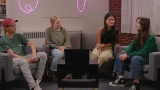 Four people sit on chairs around a table with a laptop in a cozy room. A pink neon light decorates the brick wall, creating a relaxed vibe.