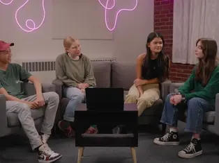 Four people sit on chairs around a table with a laptop in a cozy room. A pink neon light decorates the brick wall, creating a relaxed vibe.