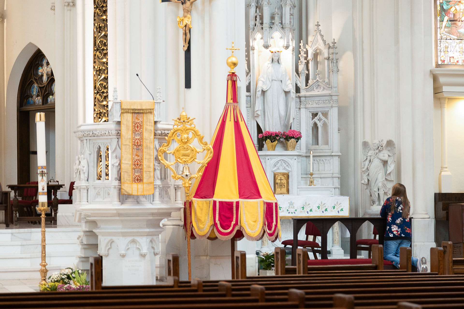 Church interior with a woman kneeling in prayer. Ornate altar with yellow and red canopy, statue, and floral decorations. Peaceful atmosphere.