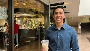 Smiling man in a blue shirt holds iced coffee outside a café with "More Coffee" sign. Warm lighting and wooden floors in the background.
