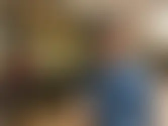 Smiling man in a blue shirt holds iced coffee outside a café with "More Coffee" sign. Warm lighting and wooden floors in the background.