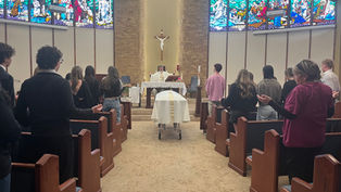 People in a chapel attend a service led by a priest. A casket draped in white is in the center. Stained-glass windows in the background.