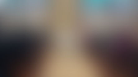 People in a chapel attend a service led by a priest. A casket draped in white is in the center. Stained-glass windows in the background.