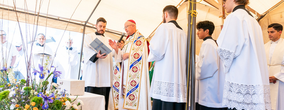 Bishop speaks with microphone to altar servers in white robes under a tent, beside a flower-filled altar in a solemn ceremony
