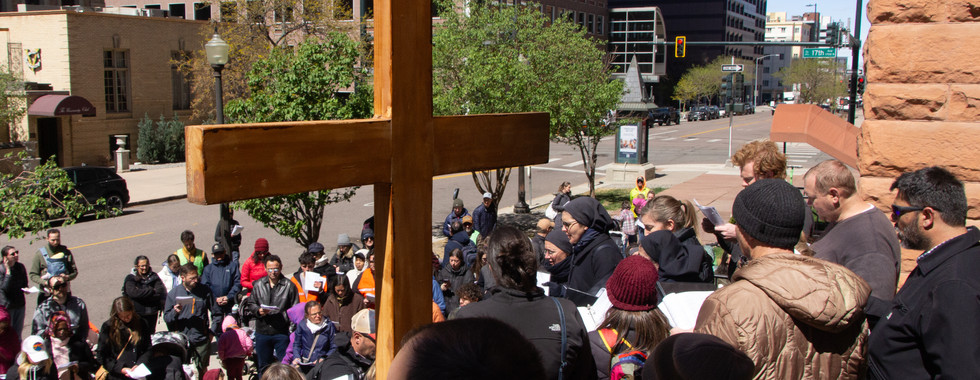 A crowd gathers in a small urban park around a statue while holding prayer sheets during a public devotion.