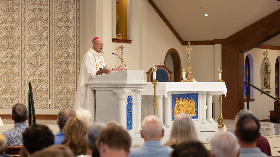 Catholic bishop in white vestments speaks at marble altar to seated congregation in a bright church with candles and gold cross.