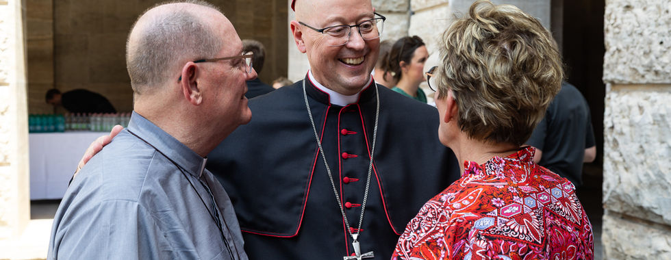 Smiling bishop in black and magenta robes chats with two people in a stone courtyard.