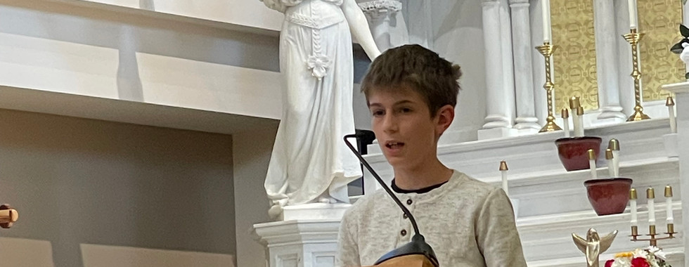 Boy speaking at a church lectern with an open book. A white statue of an angel with a trumpet is in the background. Formal setting.