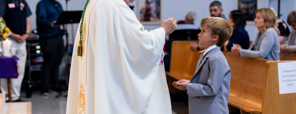 Priest in white robe giving communion to boy in gray suit in a church. Congregation seated, banners visible. Ceremony atmosphere.