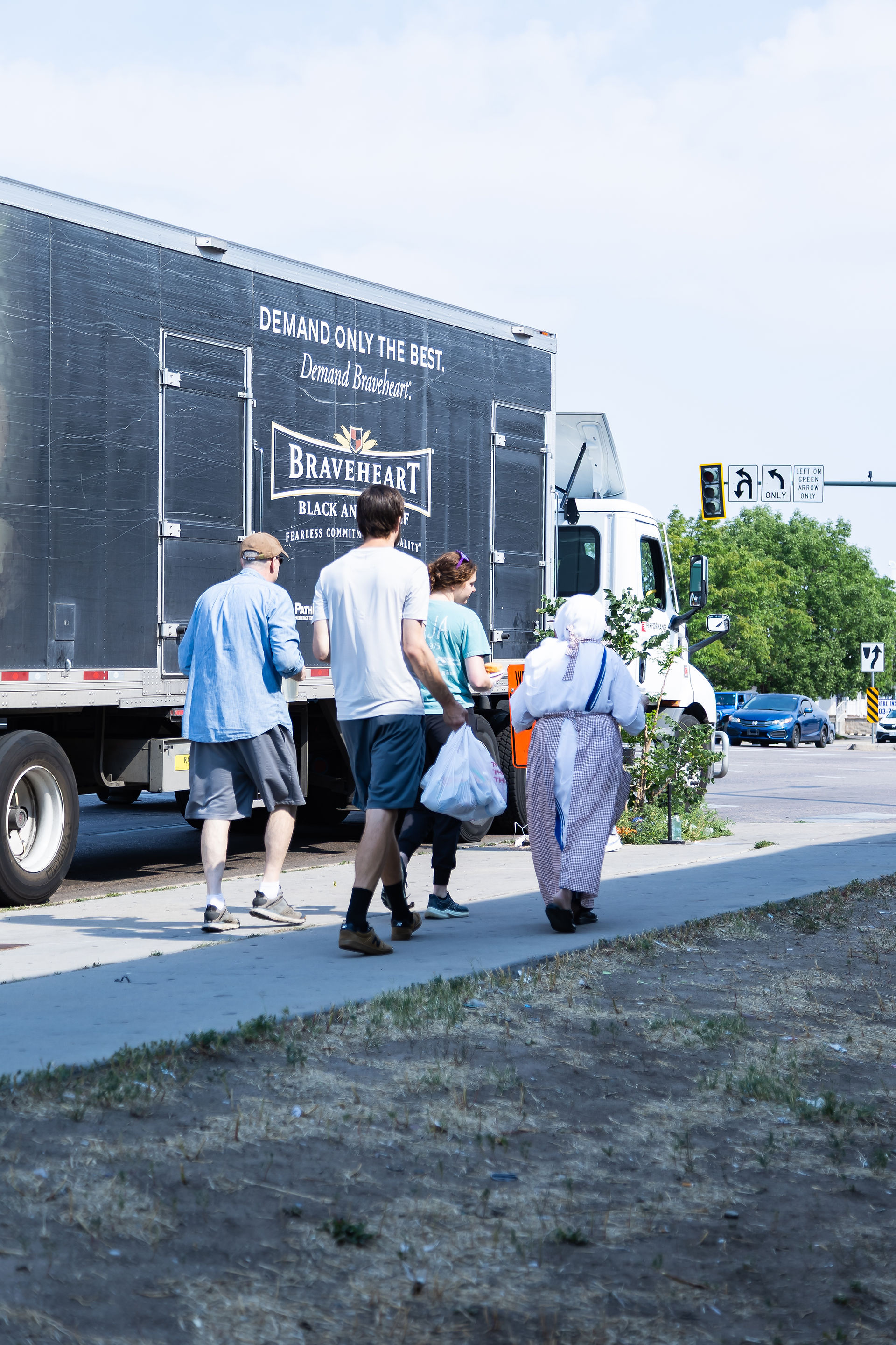 Four people walk along a sidewalk beside a parked truck with Braveheart branding. The setting is a sunny street with traffic signs.
