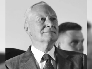 Elderly man in a suit gazes upward with a thoughtful expression. Blurred figure in the background. Black and white image.
