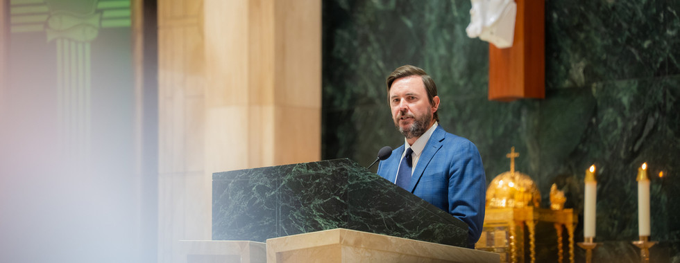 Man in a blue suit speaks at a marble lectern in a church, with candles and a crucifix behind him.