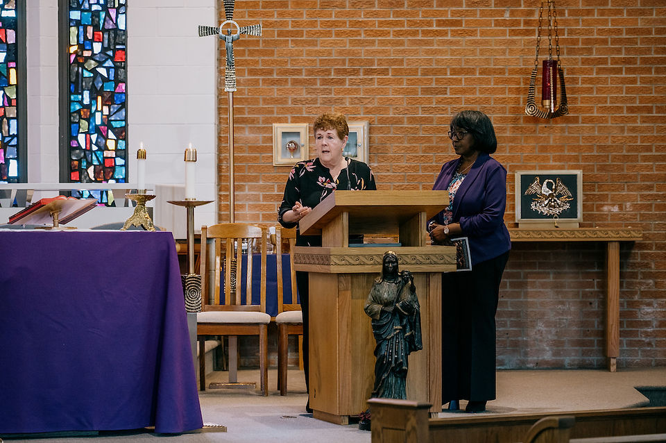 Two women stand at a wooden lectern in a church, one speaking. Stained glass window and religious decor are visible. Mood is solemn.