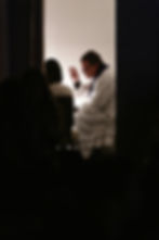 Priest in white robe and scarf raises hand in blessing, seated beside a person, in a dimly lit room with a white wall backdrop.