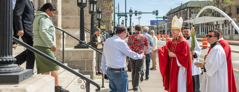 A person in red robes greets pedestrians on a sunny city street. Others descend stone steps. Orange cones and traffic signs in background.