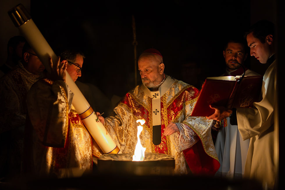 Bishop in gold and red vestments blesses a fire during the Easter Vigil, as clergy assist and hold a large Paschal candle and a liturgical book.