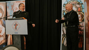 Two clergy members laughing on stage, one at a podium with a cross emblem. Background shows religious artwork, creating a joyful mood.