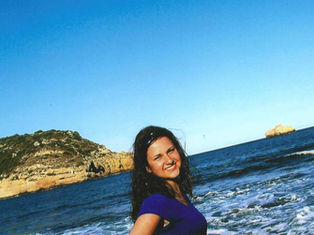 Smiling woman in a blue shirt stands by the ocean, with waves and rocky cliffs in the background under a clear blue sky.