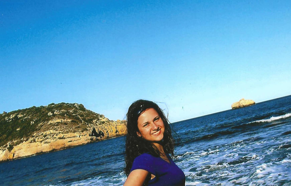 Smiling woman in a blue shirt stands by the ocean, with waves and rocky cliffs in the background under a clear blue sky.