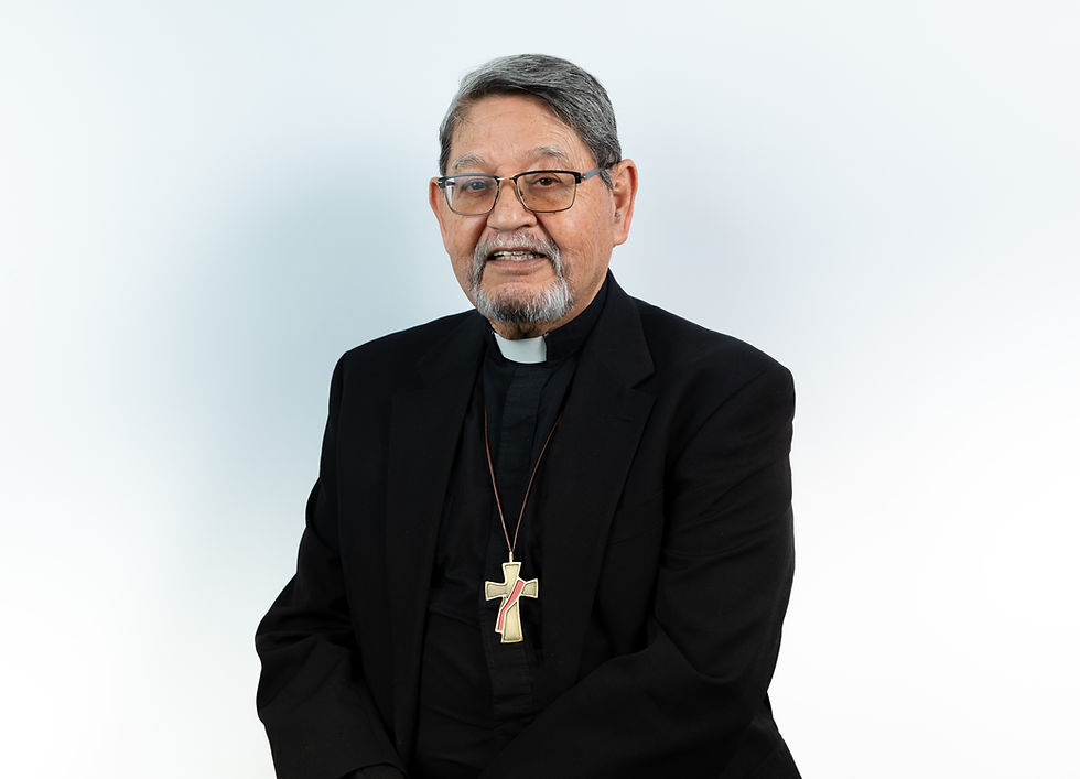 Elderly man in a black suit with clerical collar, wearing glasses and a cross necklace, smiling against a plain white background.