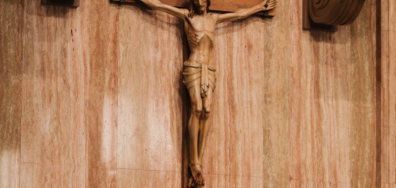 Crucifix on a marble church altar with candles, red flowers, and gold tabernacle; Latin arch text above creates a solemn mood