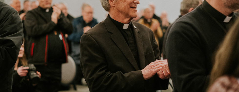 Elderly man in clerical attire claps in a conference room, surrounded by other clapping attendees. Table with papers and a water bottle in foreground.