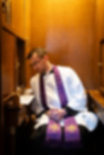 A priest in a wooden confessional dressed in a white alb with a purple stole around his neck.